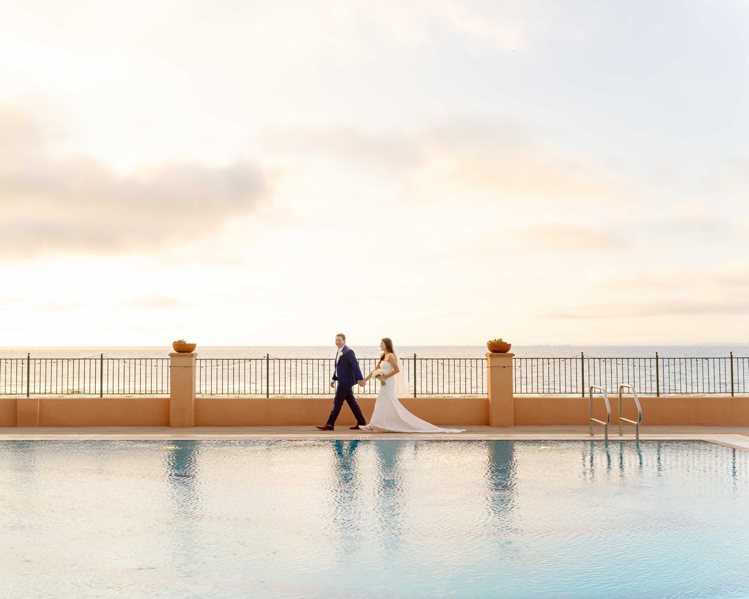 bride and groom holding hands walking along large blue pool at sunset