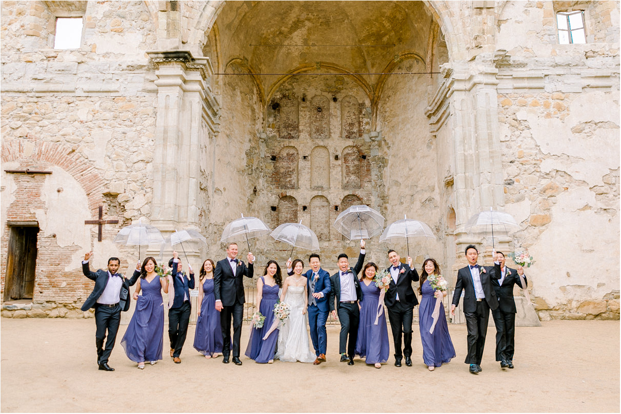 Large wedding party in black suits and purple dresses walking towards the camera holding clear umbrellas