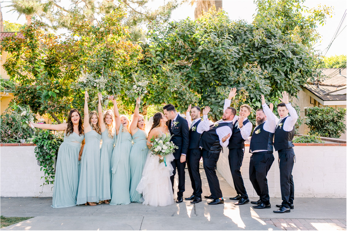 Bride and groom with wedding party celebrating as they kiss on sidewalk