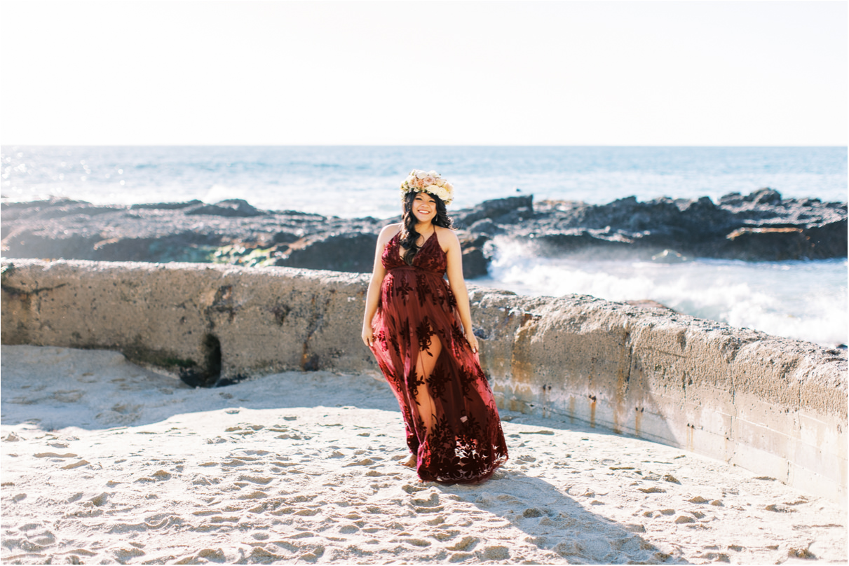 Pregnant woman in maroon dress and rose flower crown walking on sand towards camera with beach behind her