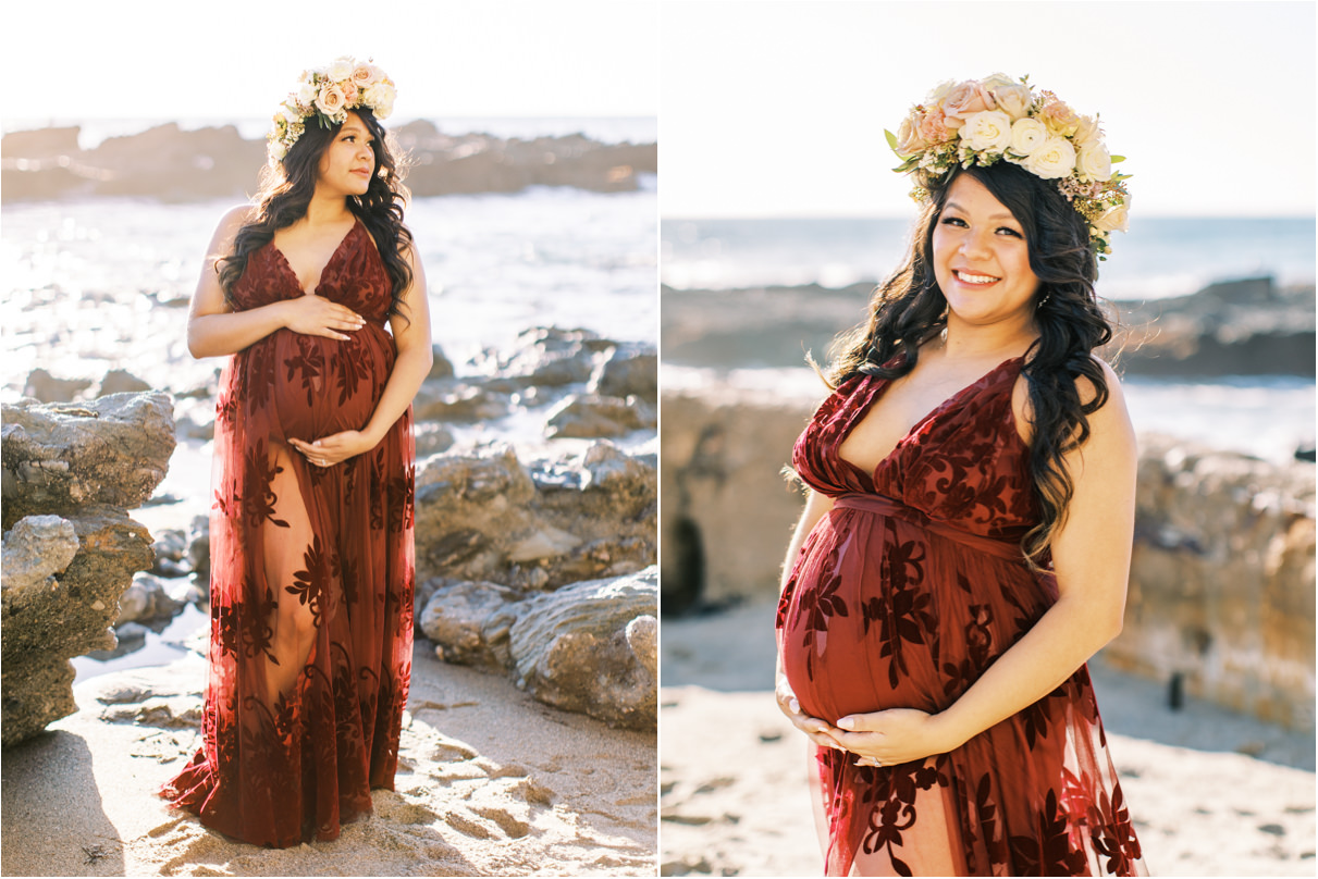 Pregnant woman in maroon dress and rose flower crown posing on sand at the beach holding her belly