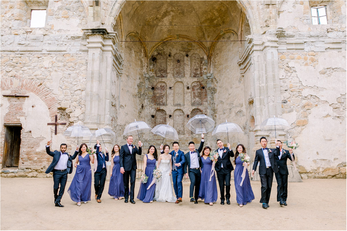 Bride and groom with entire wedding party using clear umbrellas for rain and Mission San Juan Capistrano