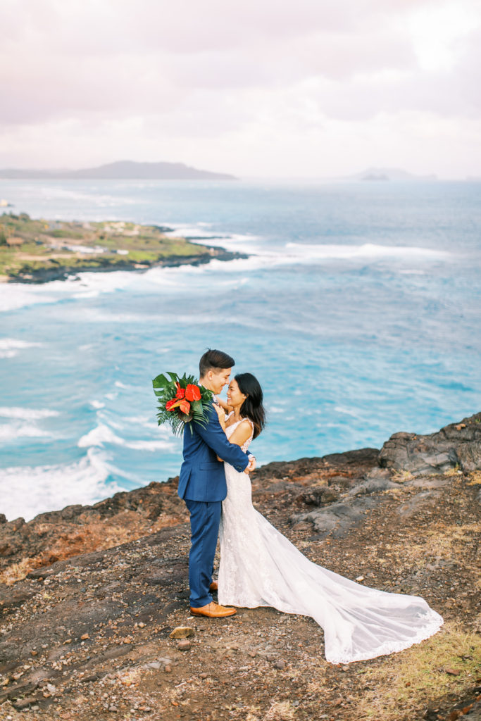 Bride and groom facing each other on ocean lookout with blue water beneath them on oahu