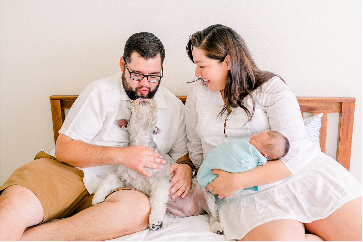 Parents are sitting on bed holding newborn baby and dad is kissing dog